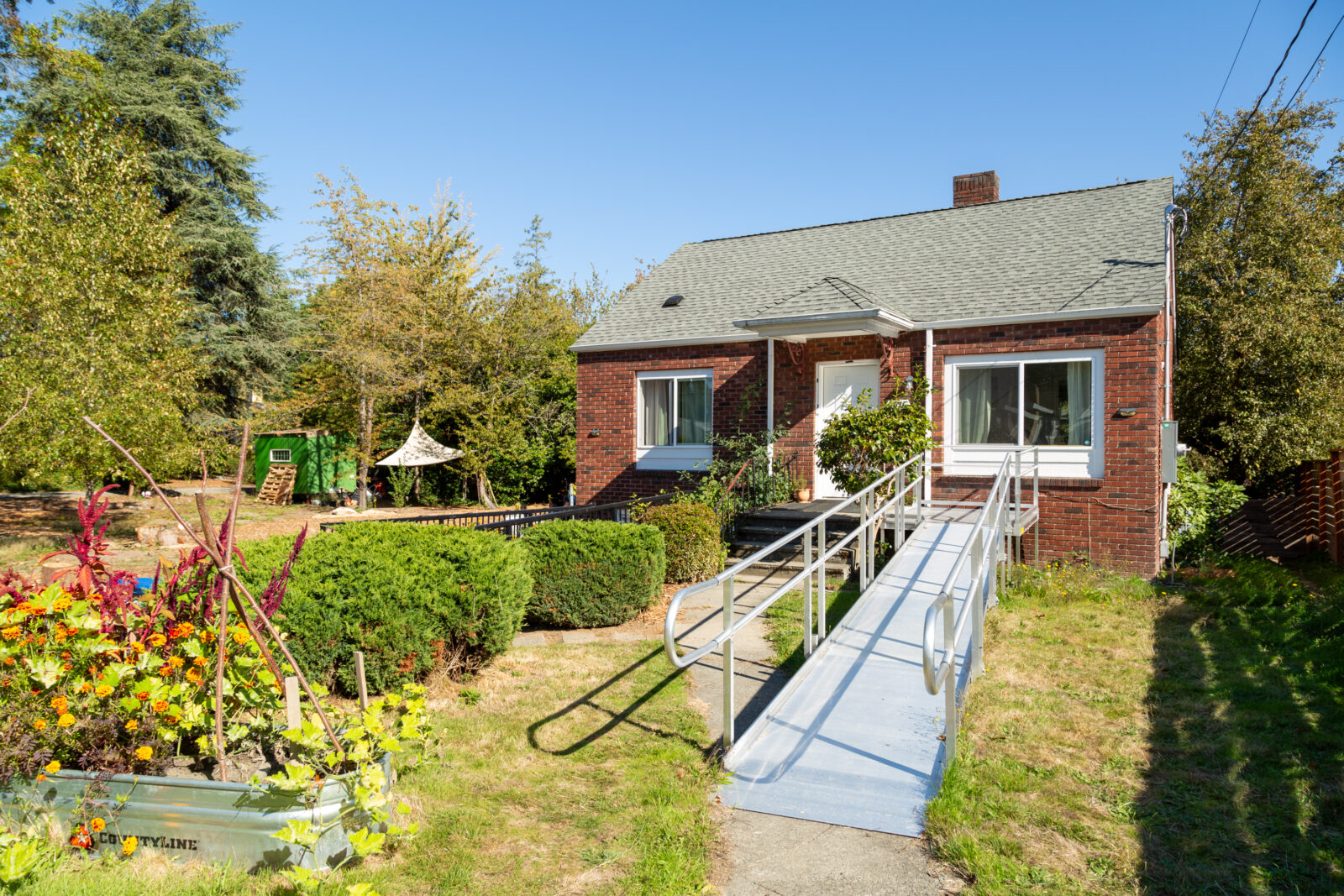 A small brick house with a sloped gray roof and a wooden ramp leading to the front door. The yard features colorful flowers, shrubs, and a play structure. It is a clear, sunny day with a blue sky.